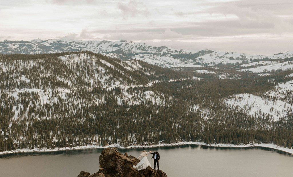 wedding couple hiking down a rock with snow covered mountains and a lake behind them
