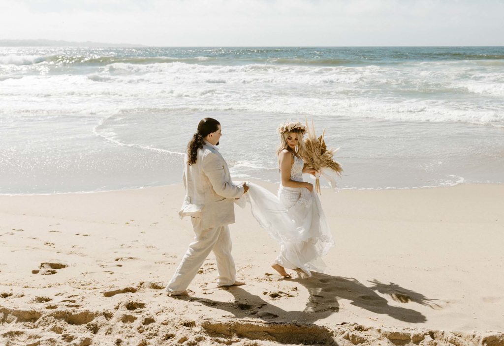 groom holding brides dress as they walk on the beach in monterey