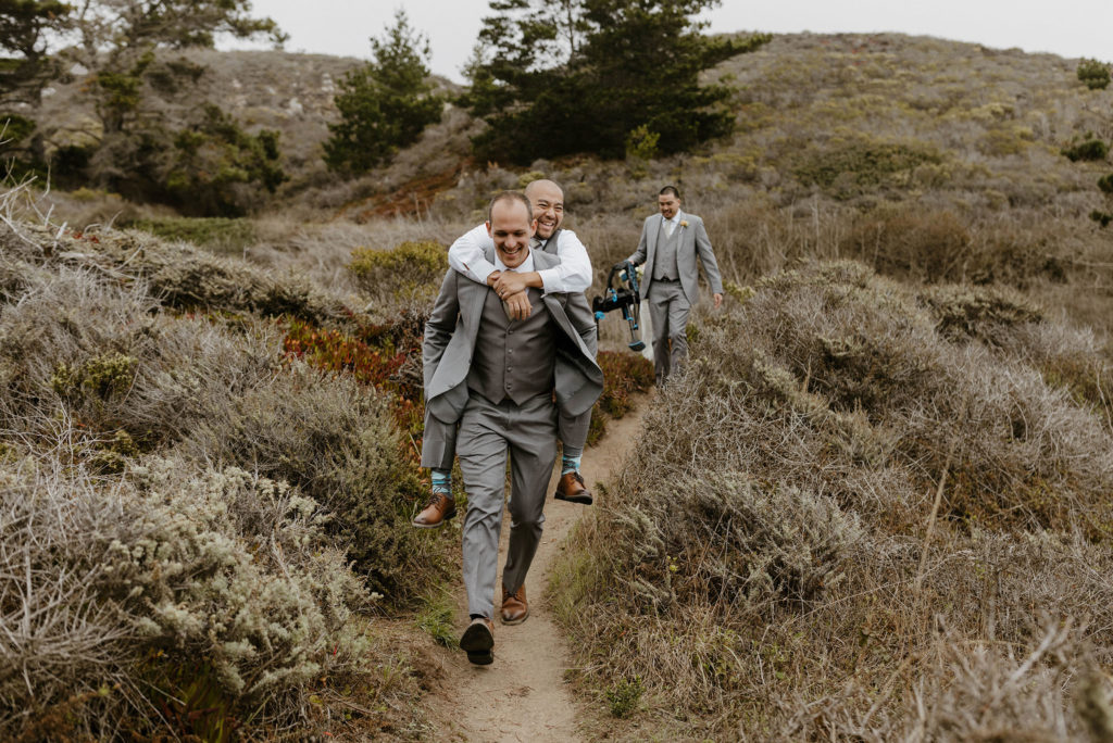 Groomsman laughing while giving another groomsman piggyback ride while walking along trail at carmel highlands