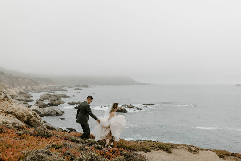 Wedding groom holding brides wedding dress as they walk down trail along rocky cliffs next to water at carmel highlands