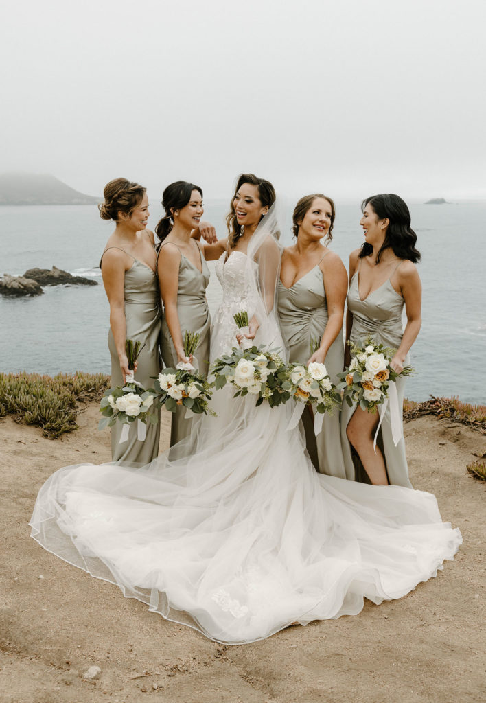 Wedding bride smiling while standing with bridesmaids on rocky shoreline with lake in background at carmel highlands 