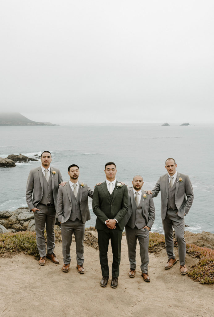 Wedding groom standing with hands clasped while groomsmen stand beside him with arms on shoulders at carmel highlands