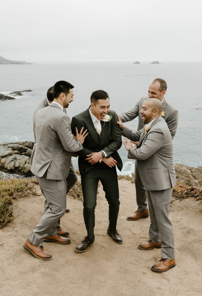 Wedding groom laughing while groomsmen play shove him while standing on rocky shoreline with lake in background at carmel highlands