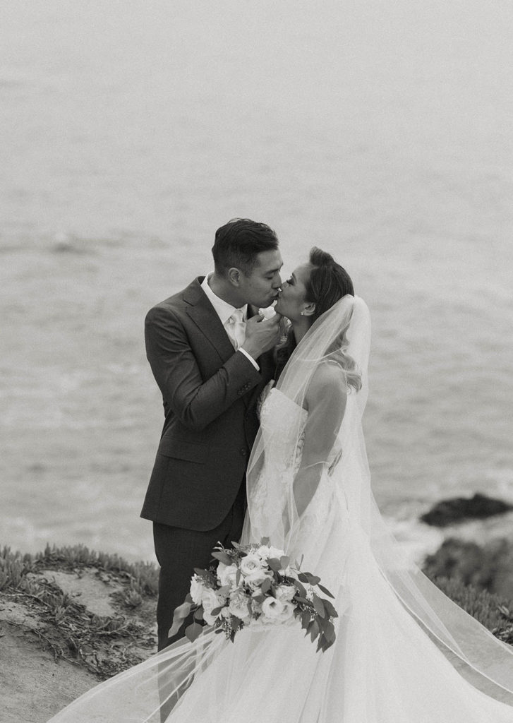 Wedding couple kissing while standing on rocky shoreline at carmel highlands