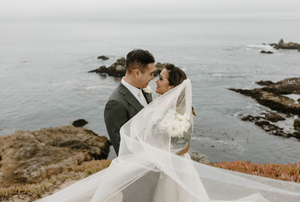 Wedding couple smiling while holding each other on a cliff with lake in background at carmel highlands