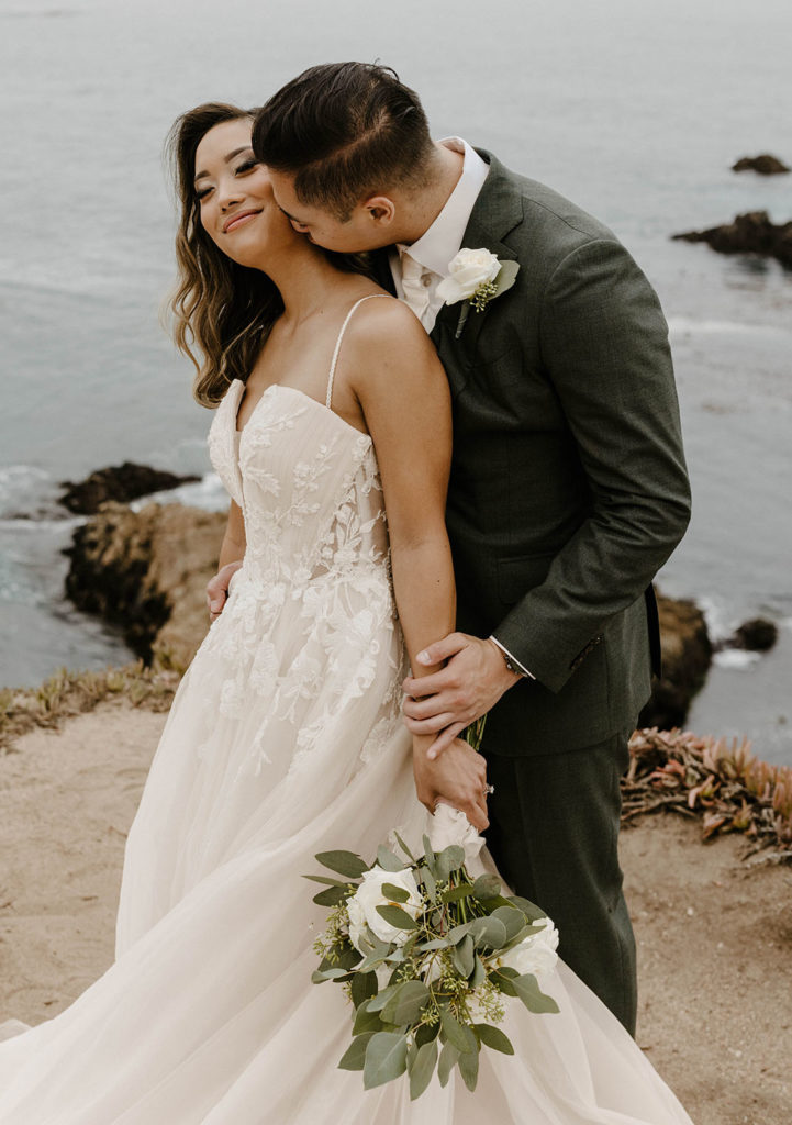 Wedding groom kissing bride on neck as she smiles while on rocky shoreline at carmel highlands with lake in background
