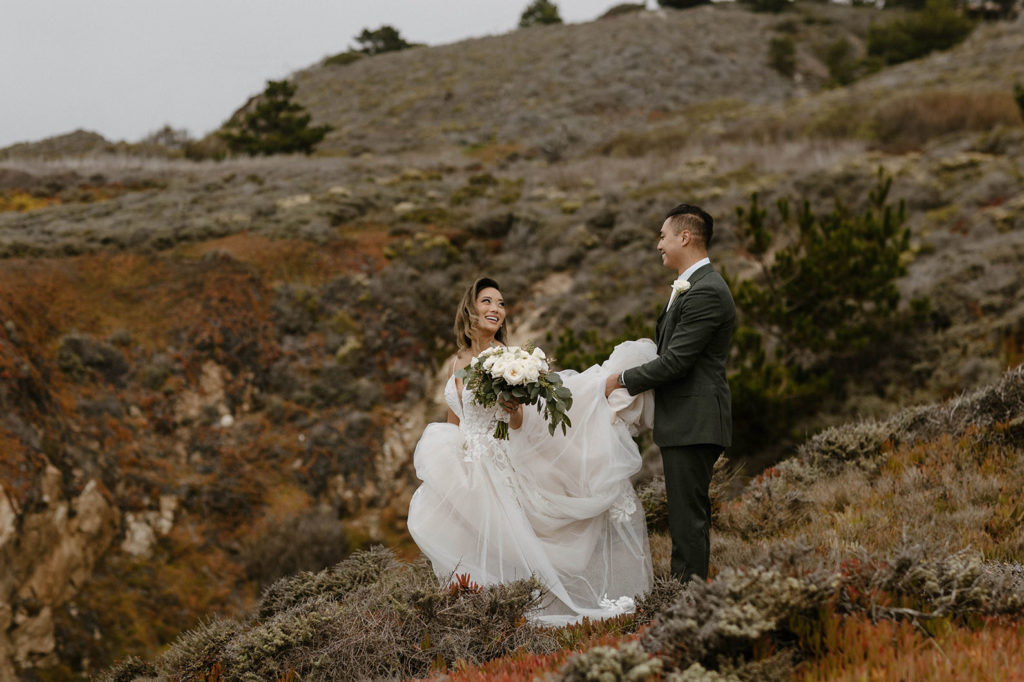 Wedding bride smiling back at groom as he holds her wedding dress up while walking along trail at carmel highlands