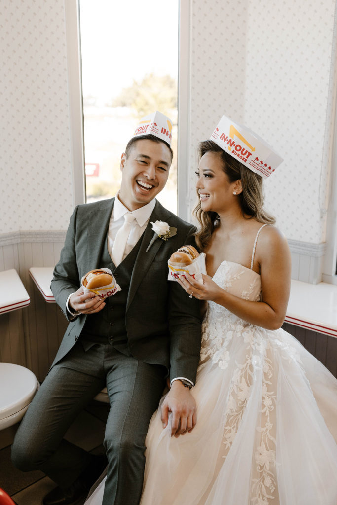 Wedding couple sitting together holding burgers and wearing paper hats inside at restaurant
