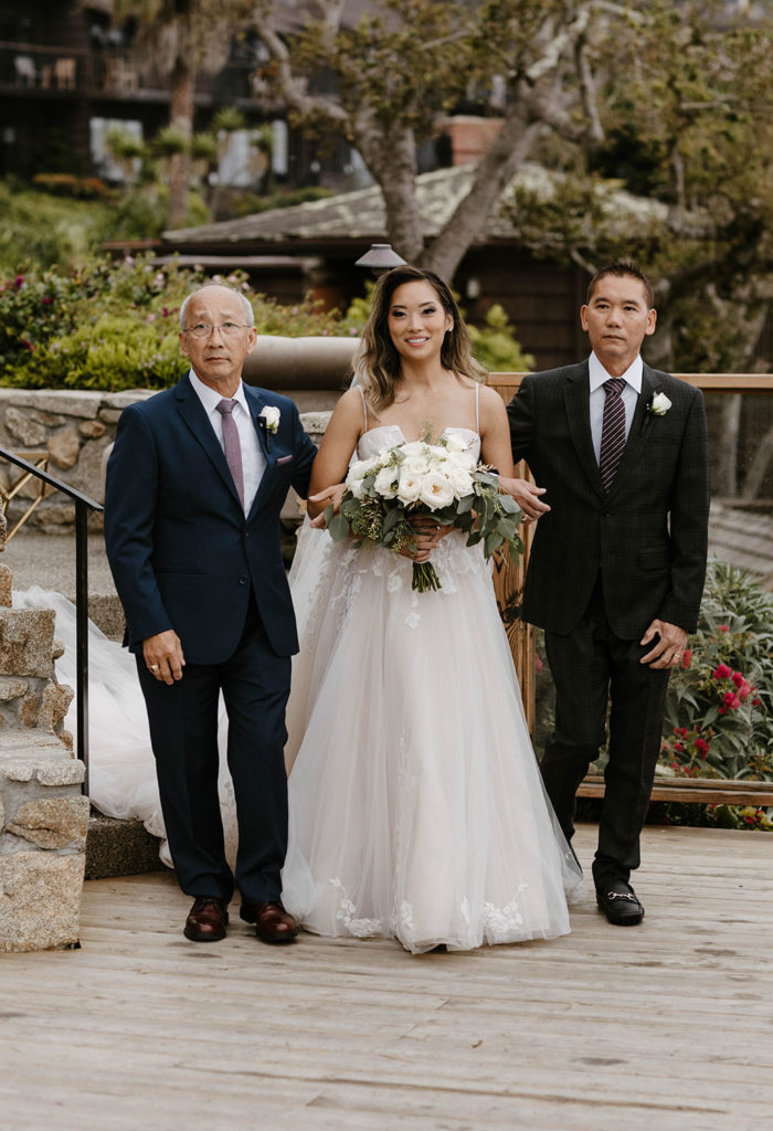 Wedding bride walking down wedding aisle as dads hold either arm at carmel highlands