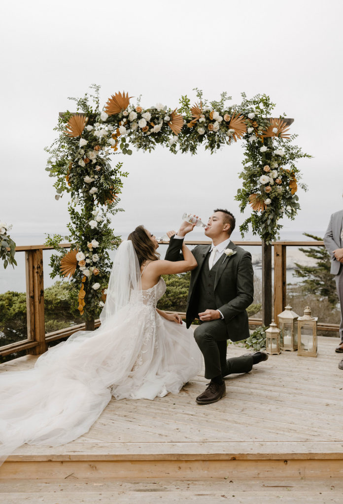 Wedding couple on one knee together chugging drinks during wedding ceremony at carmel highlands