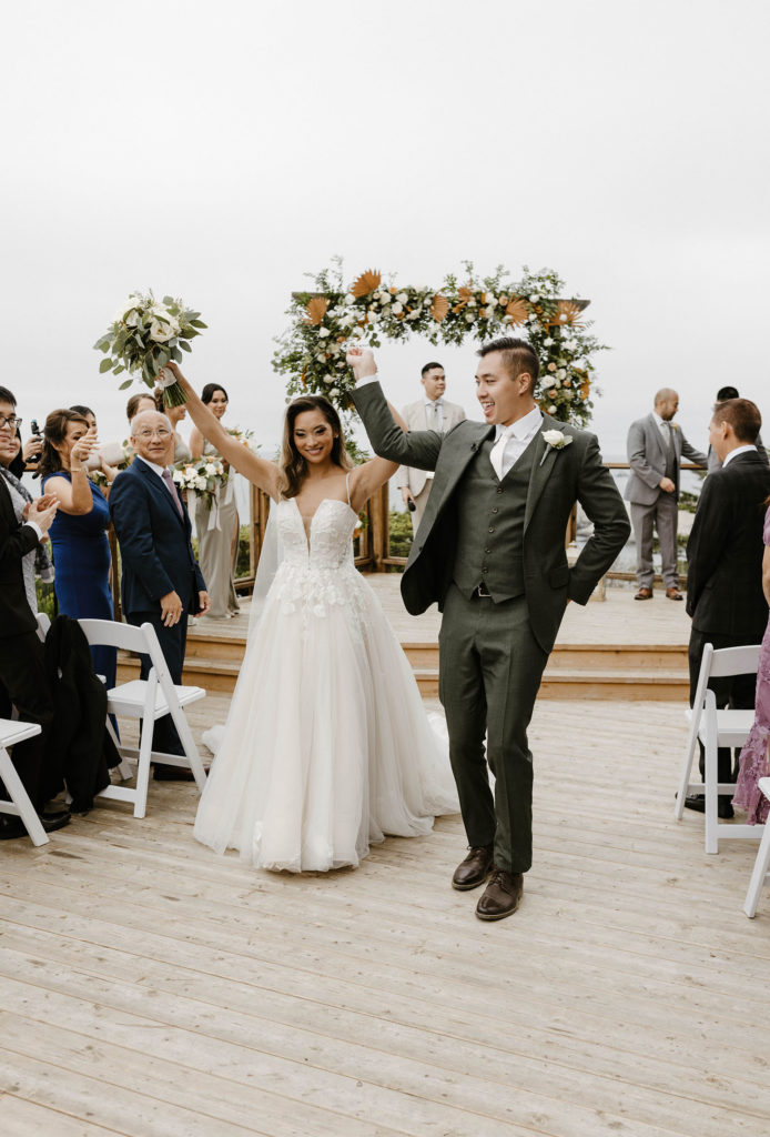 Wedding couple holding hands in the air and celebrating while walking down aisle together after ceremony at carmel highlands