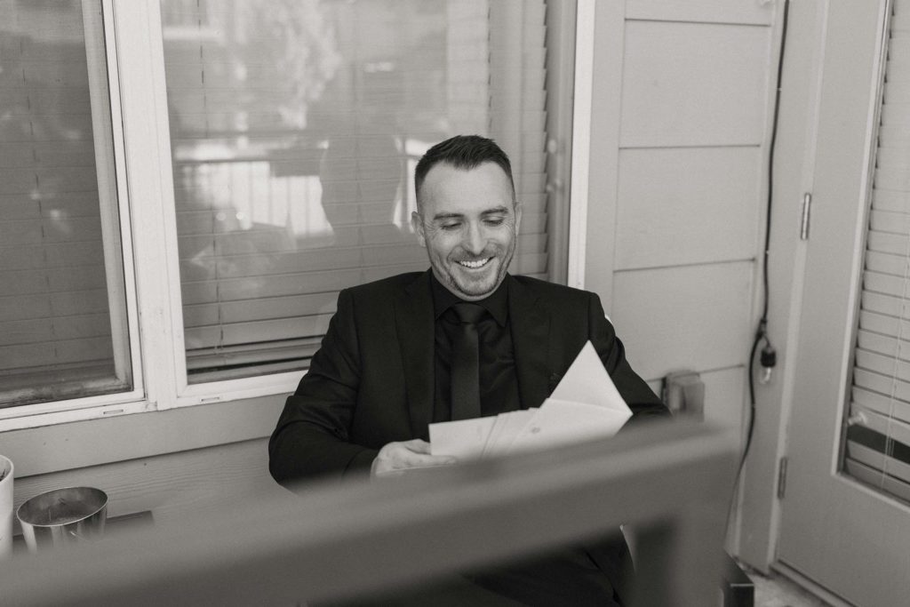Wedding groom sitting in a chair reading letters while smiling outside at the Bell Tower