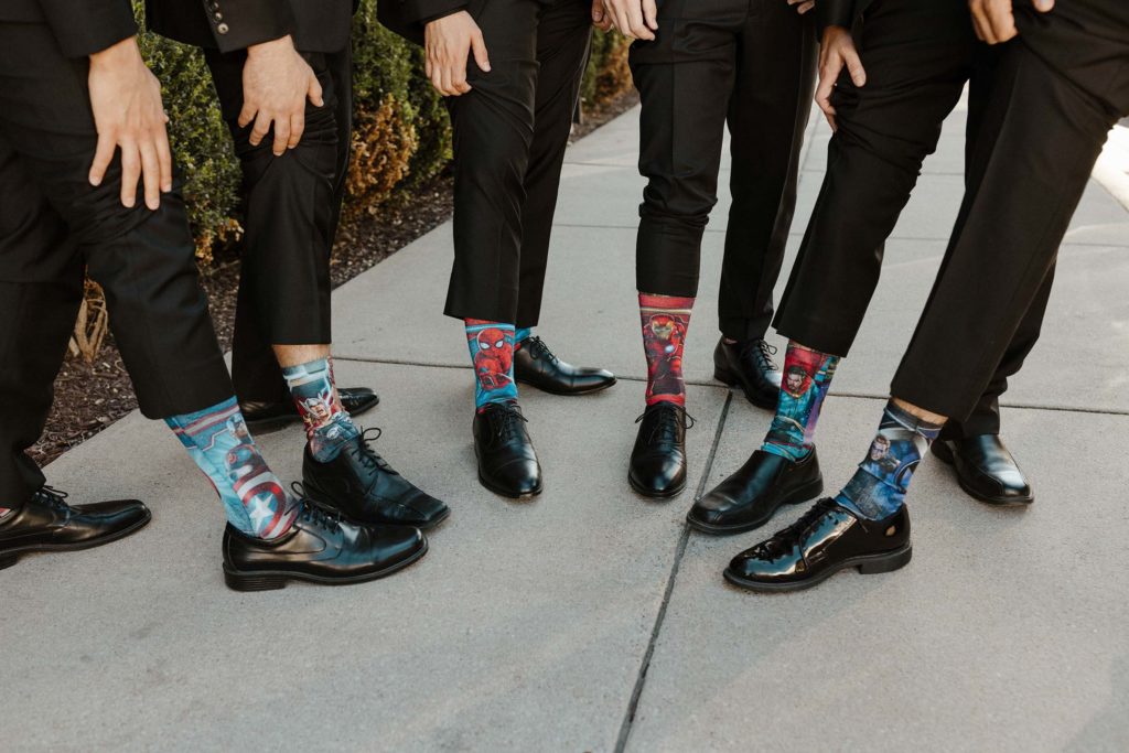 Wedding groomsmen holding feet together and lifting up pant legs to show matching super hero socks 