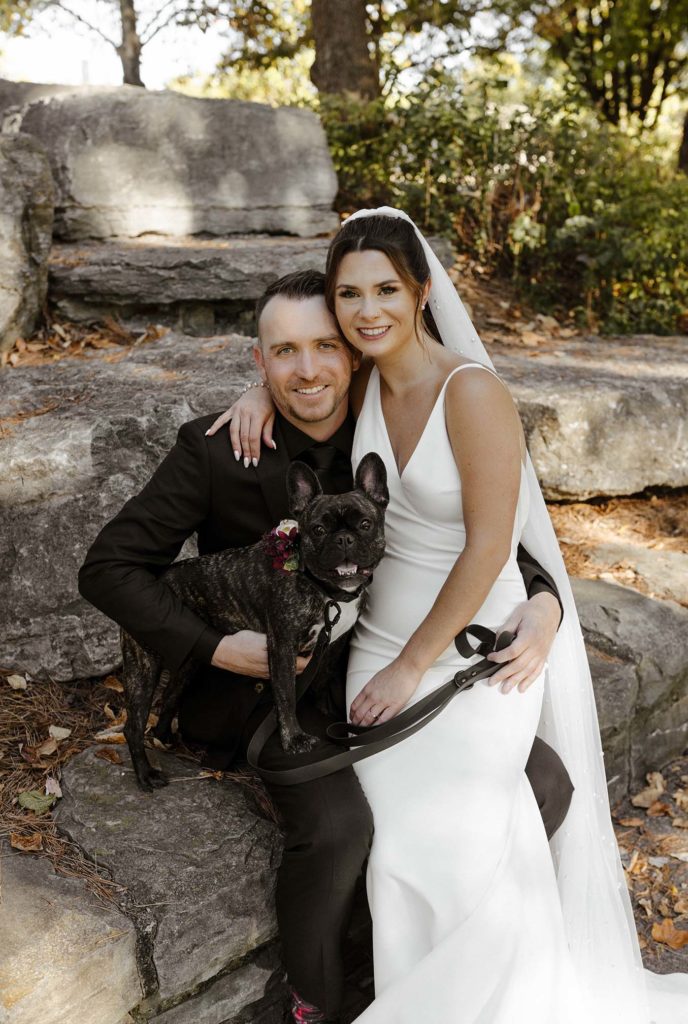 Wedding couple sitting on rock together while holding dog while all smile at camera outside at the Bell Tower