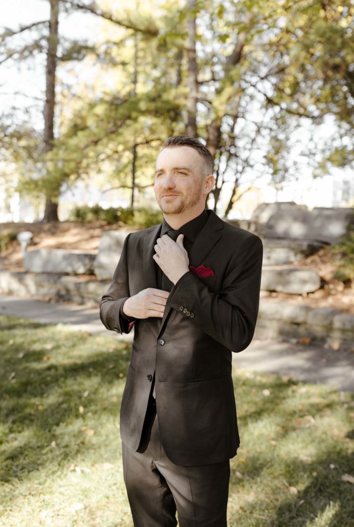 Wedding groom smiling while looking into distance and adjusting tie while outside at the Bell Tower