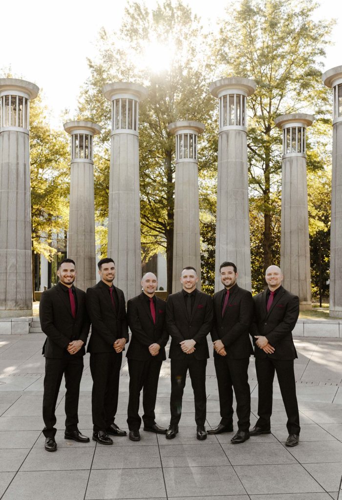 Wedding groom with groomsmen standing in front of large columns with hands clasped and smiling at camera