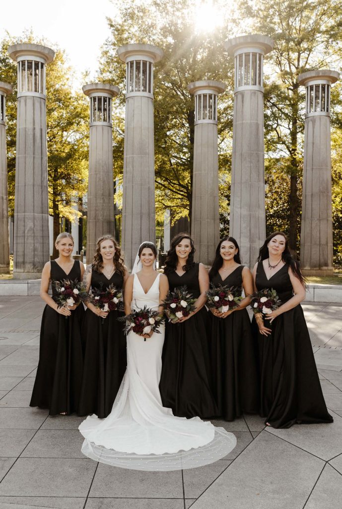 Wedding bride and bridesmaids standing together holding flower bouquets while smiling at camera in front of large columns