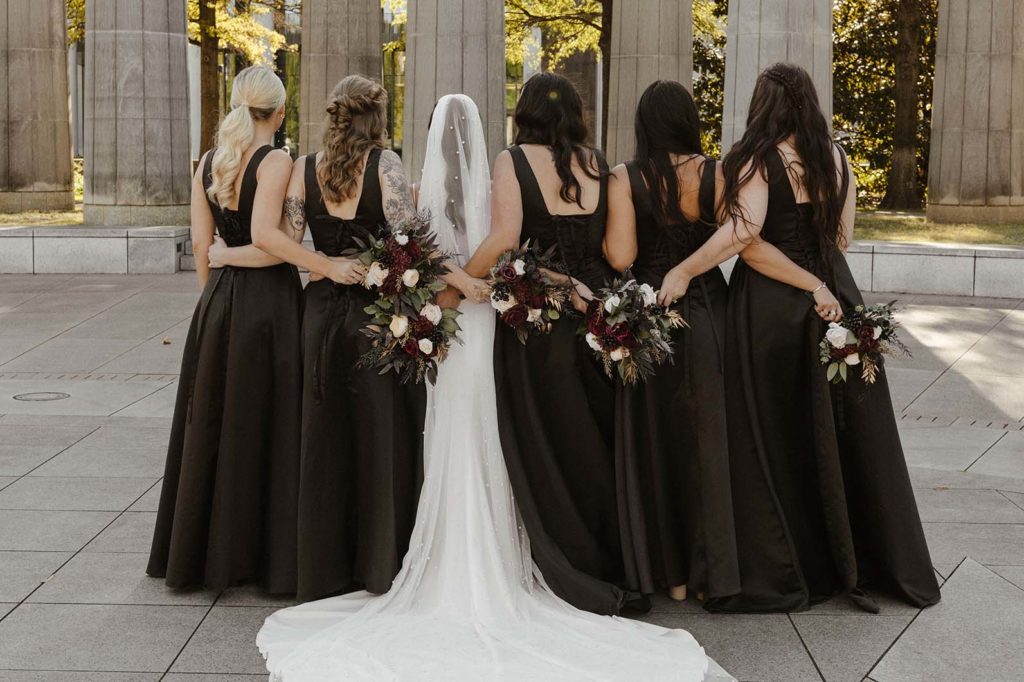 Wedding bride and bridesmaids with arms around each other holding flower bouquets while standing outside at the Bell Tower