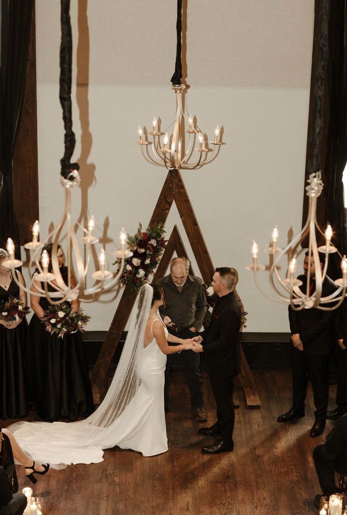 Wedding couple holding hands looking at officiant with hanging lights inside at the Bell Tower