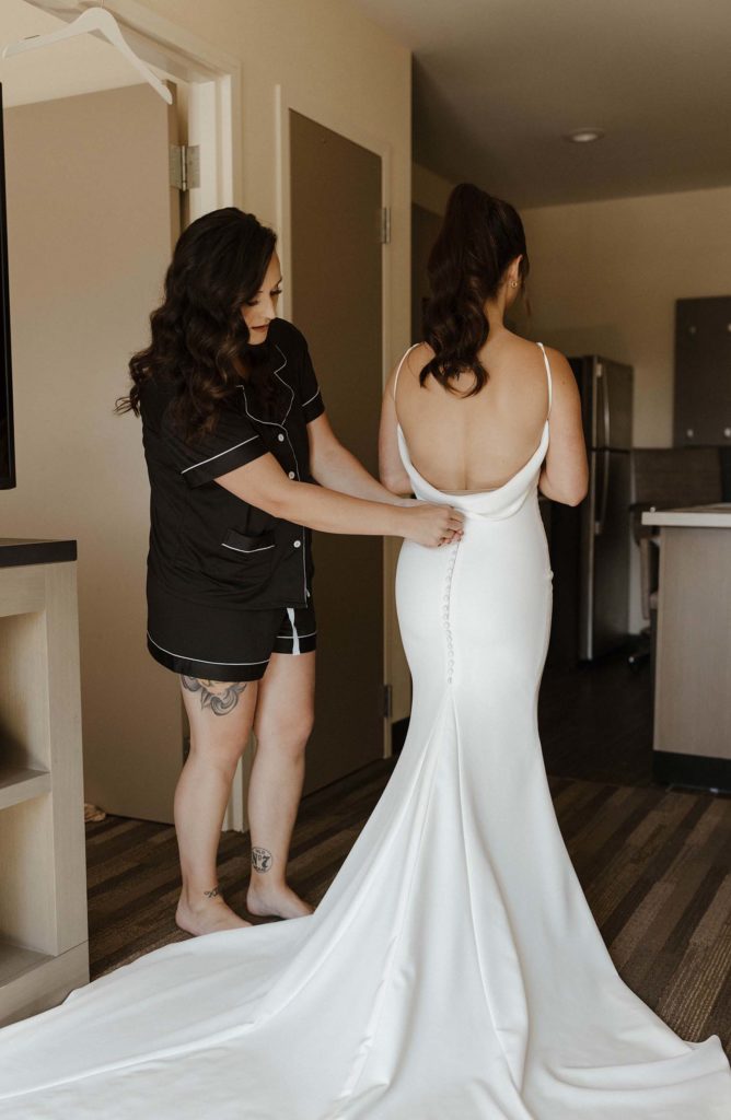 Bridesmaid helping button up the back of brides wedding dress while inside at the Bell Tower