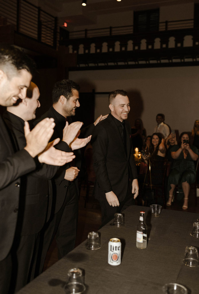 Groomsmen patting groom on back while clapping and laughing after losing at flip cup during reception at the Bell Tower