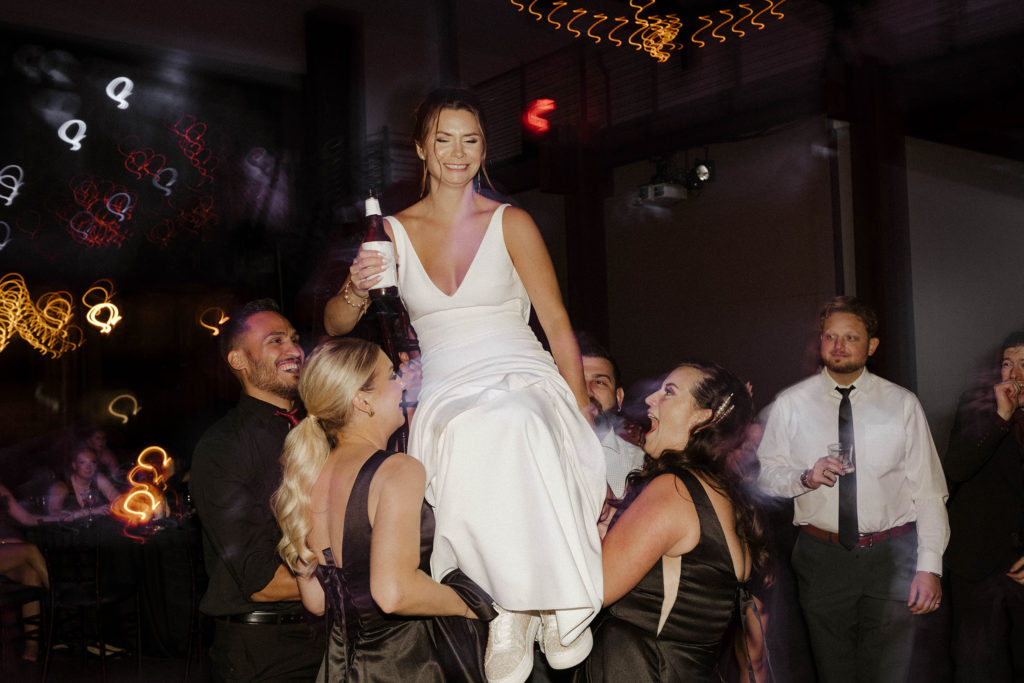 Wedding bride being lifted up on chair while holding beer and grimacing during reception at the Bell tower