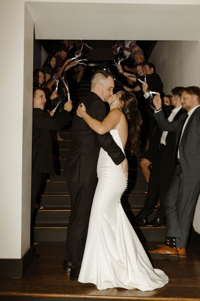 Wedding couple holding each other and kissing at bottom of staircase while guests celebrate beside them at the Bell Tower