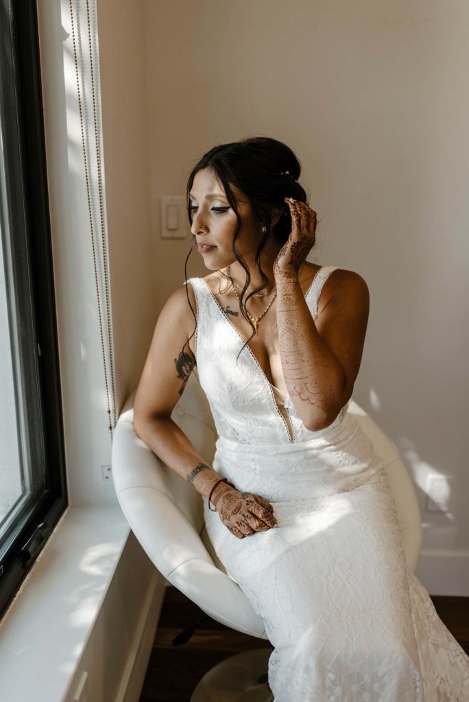 Wedding bride holding hair while sitting on white chair and looking out window at the elm estate