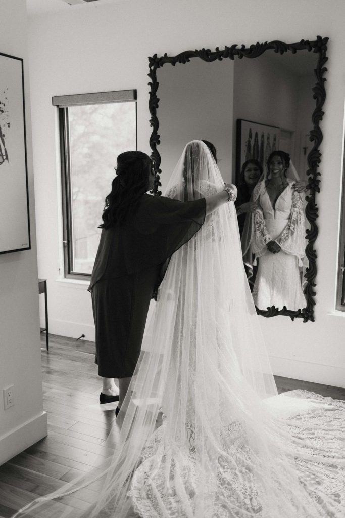 Brides mom helping wedding bride with veil while both smile in the mirror at the elm estate