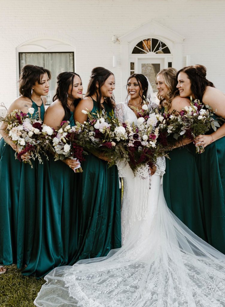 Bridesmaids smiling at wedding bride while she smiles back and all hold flower bouquets outside at the elm estate