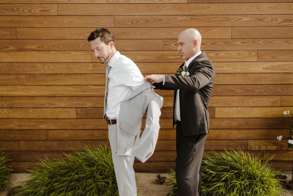 Groomsman helping wedding groom put on jacket while outside with wooden wall behind at the elm estate