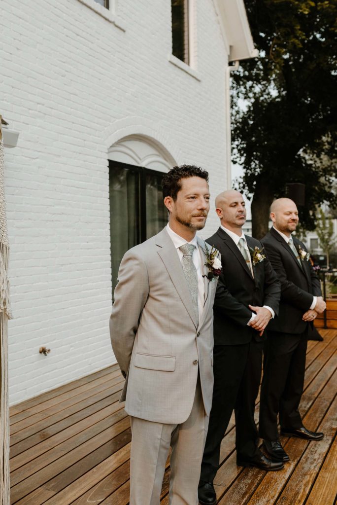 Wedding groom smiling with arms behind his back and groomsmen in background during ceremony at the elm estate