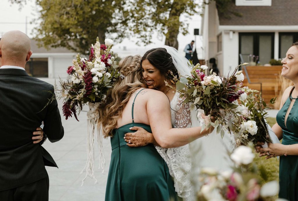 Wedding bride smiling while hugging bridesmaid and holding flower bouquet while outside at the elm estate