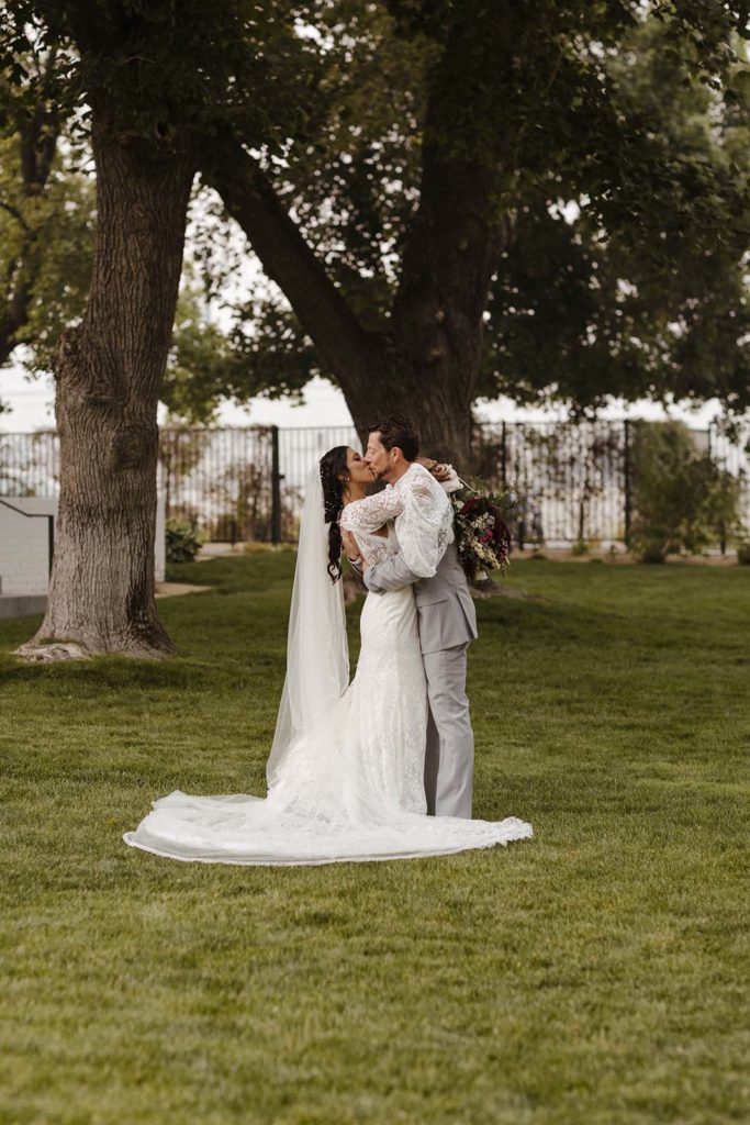 Wedding couple kissing while standing on grass with trees in background at the elm estate