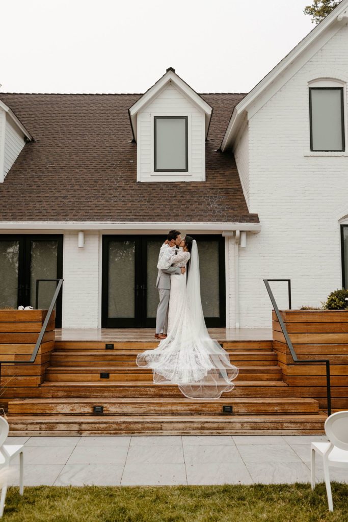 Wedding couple standing on stairs while kissing with white house in background at Elm Estate