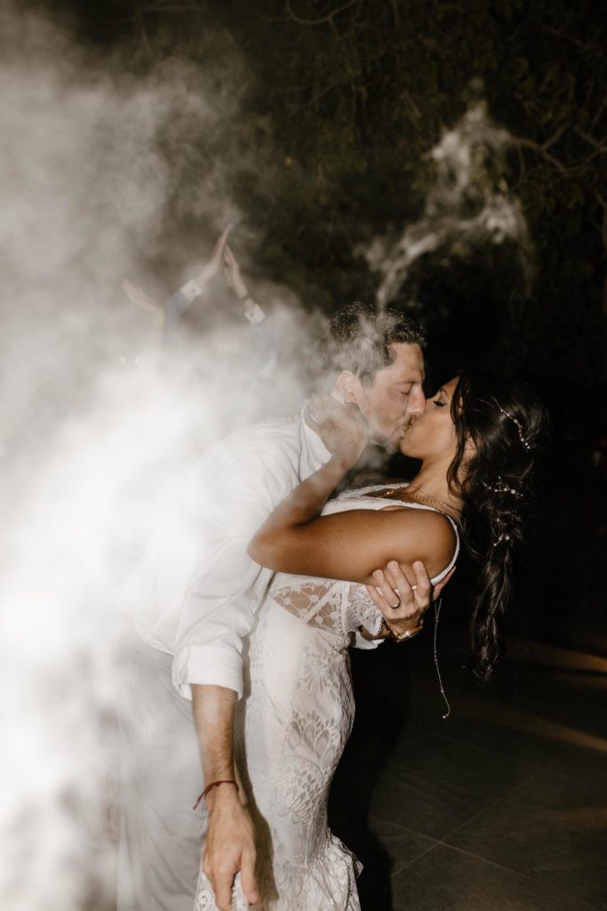 Wedding groom leaning bride back and kissing while smoke obscures them at the elm estate