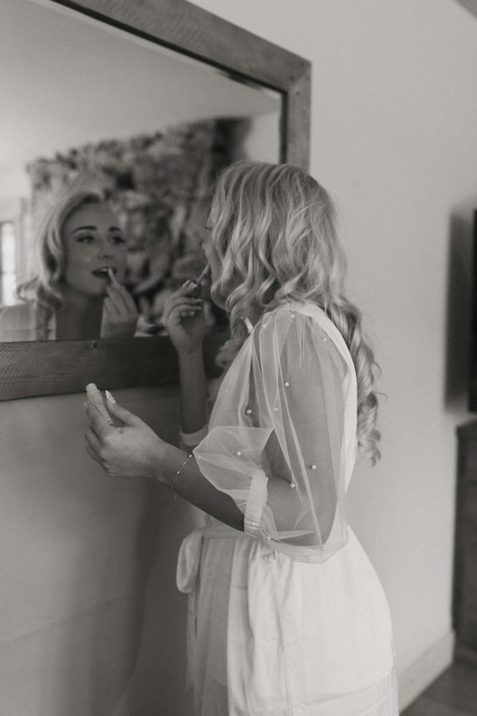 bride putting on lipstick in the mirror wearing a pearl and silk robe