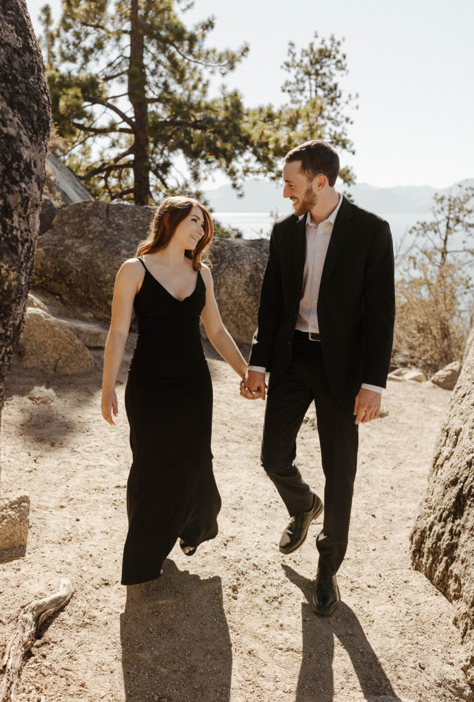 Engagement couple looking at each other and smiling while holding hands and walking along dirt trail in Lake Tahoe