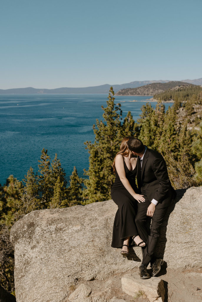 Engagement couple kissing while sitting on large rock with lake tahoe and pine trees in background