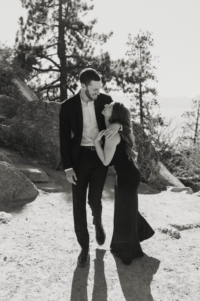 Engagement couple smiling at each other while hugging and walking along dirt trail together with large rocks in background