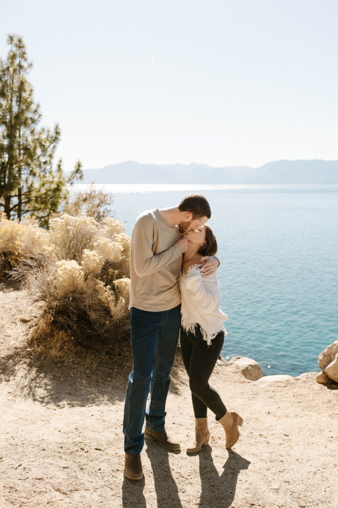 Engagement couple holding each other while standing on dirt with Lake Tahoe in background