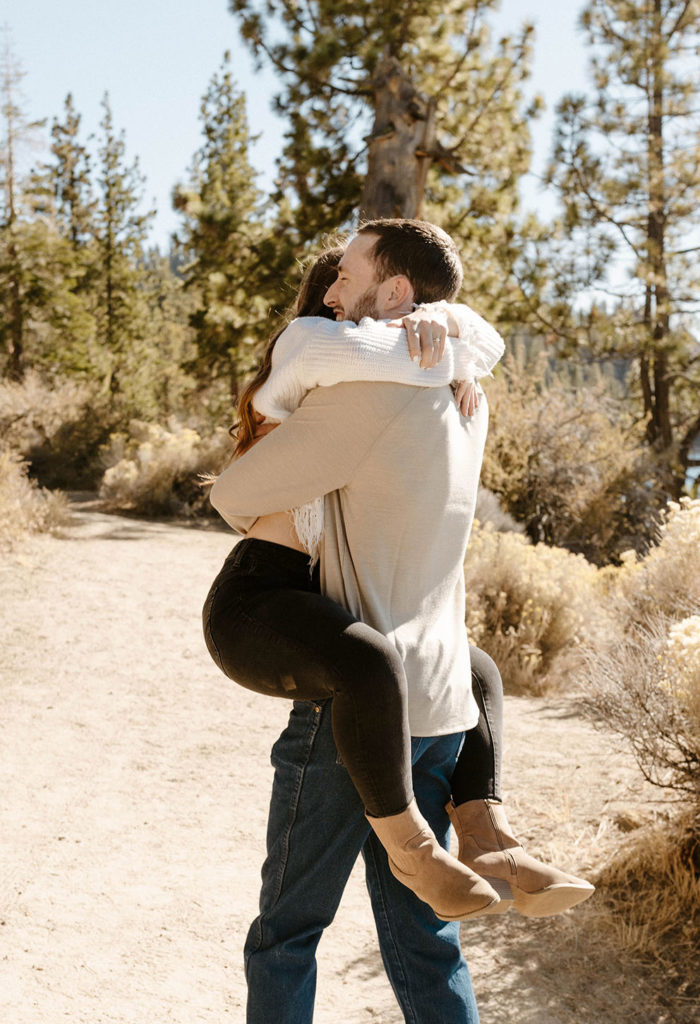 Man holding fiancé in the air while they hug on a dirt trail in Lake Tahoe