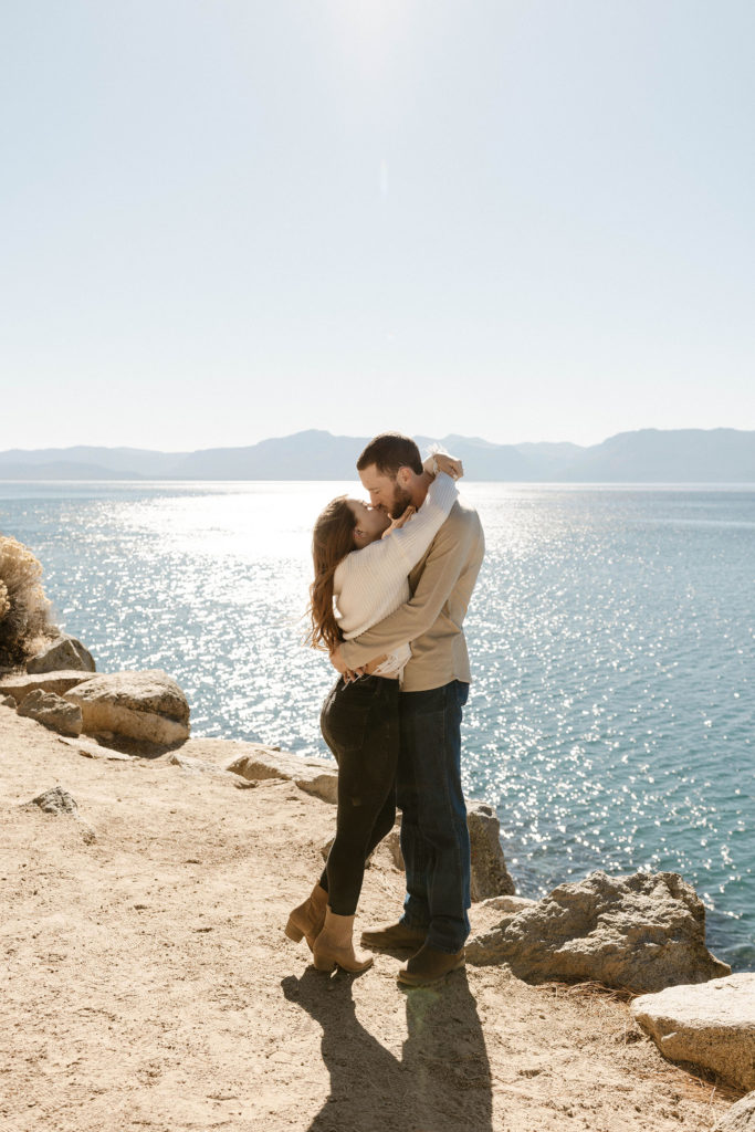 Engagement couple hugging and kissing while standing on dirt next to rocks with Lake Tahoe in background