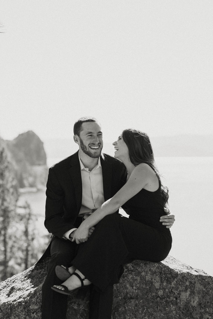 Engagement couple laughing while holding each other and sitting on rock at Lake Tahoe