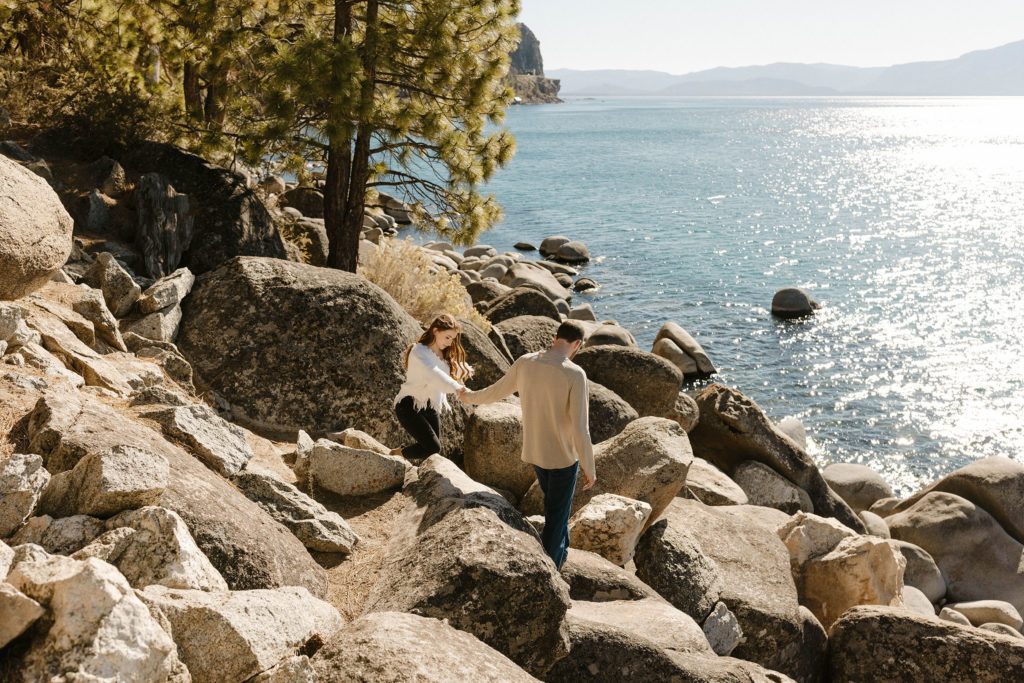 Engagement couple holding hands while walking down large rocks together toward Lake Tahoe