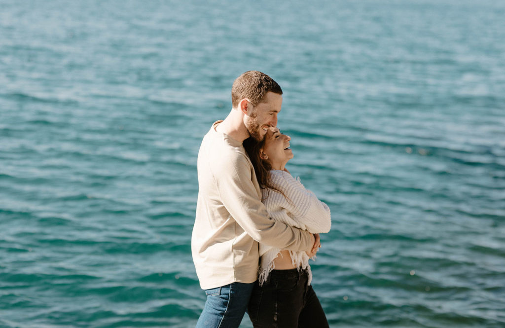 Engagement couple laughing as man hugs fiancé from behind with Lake Tahoe in background