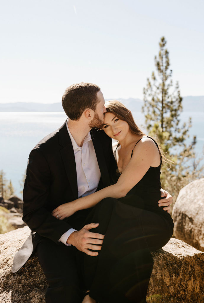 Woman looking at camera as fiancé kisses forehead while sitting on rock at Lake Tahoe