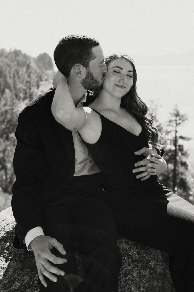 Woman holding fiancé's face as he kisses her cheek while sitting on rock at Lake Tahoe