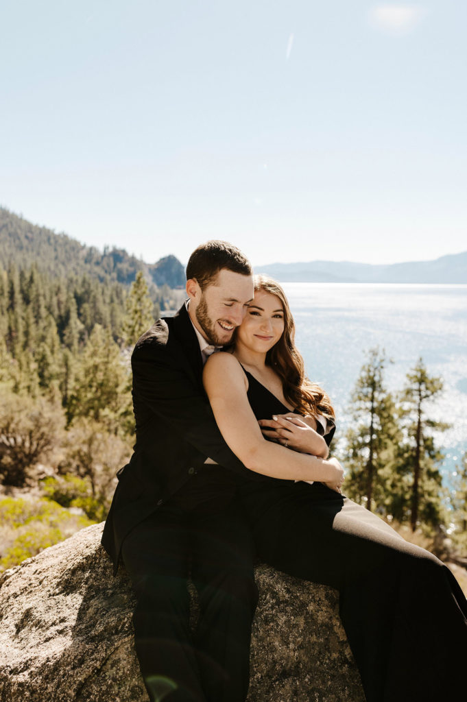 Engagement couple smiling and holding each other while sitting on rock at Lake Tahoe with pine trees in background