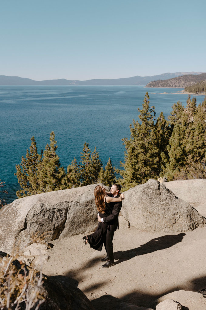 Man picking fiancé and twirling while surrounded by boulders at Lake Tahoe with lake in background